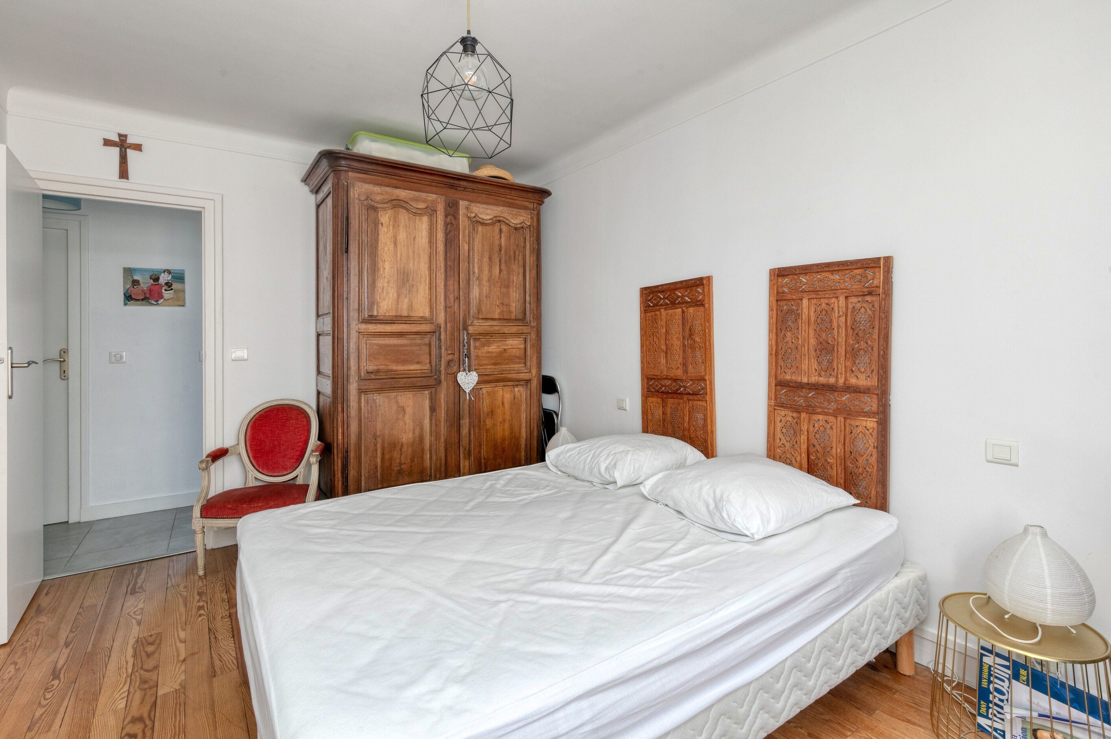 A bedroom with a bed, wooden wardrobe, a red chair, and a white lamp.