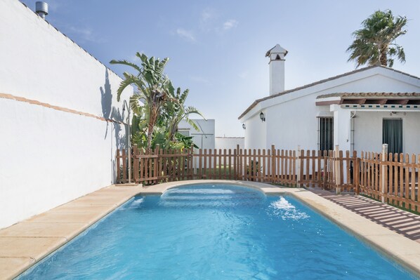 A swimming pool with a wooden deck, a white building with a red-tiled roof, and a palm tree.