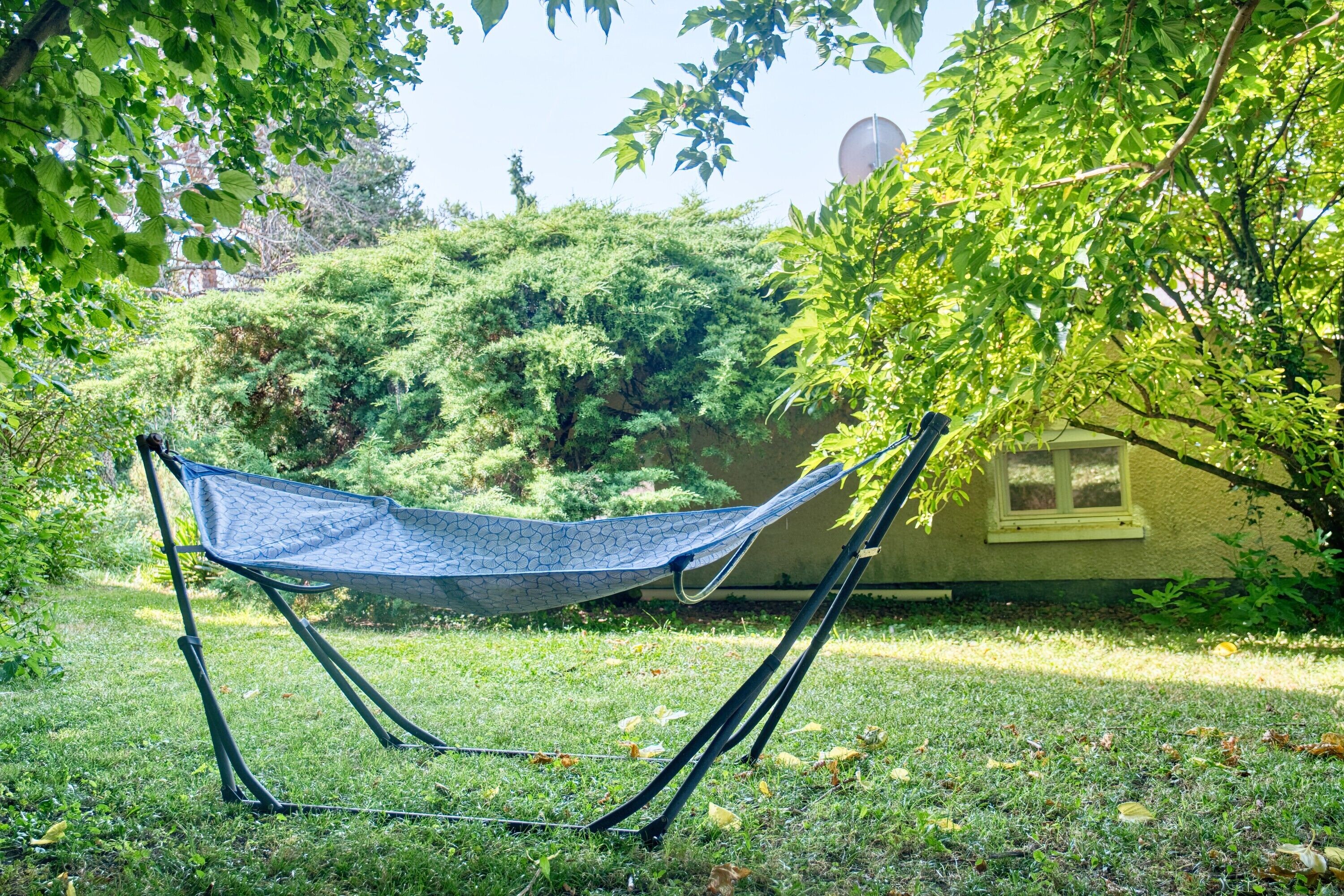 Un hamac dans une zone herbeuse, avec des arbres et un bâtiment en arrière-plan.