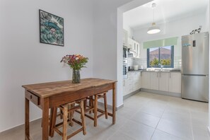A kitchen with white cabinets, a wooden table with two stools, a vase with flowers, and a refrigerator.