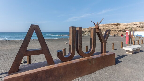 De grandes lettres métalliques épellent le mot « AJOY » sur une plage, avec un ciel dégagé et l’océan en arrière-plan.