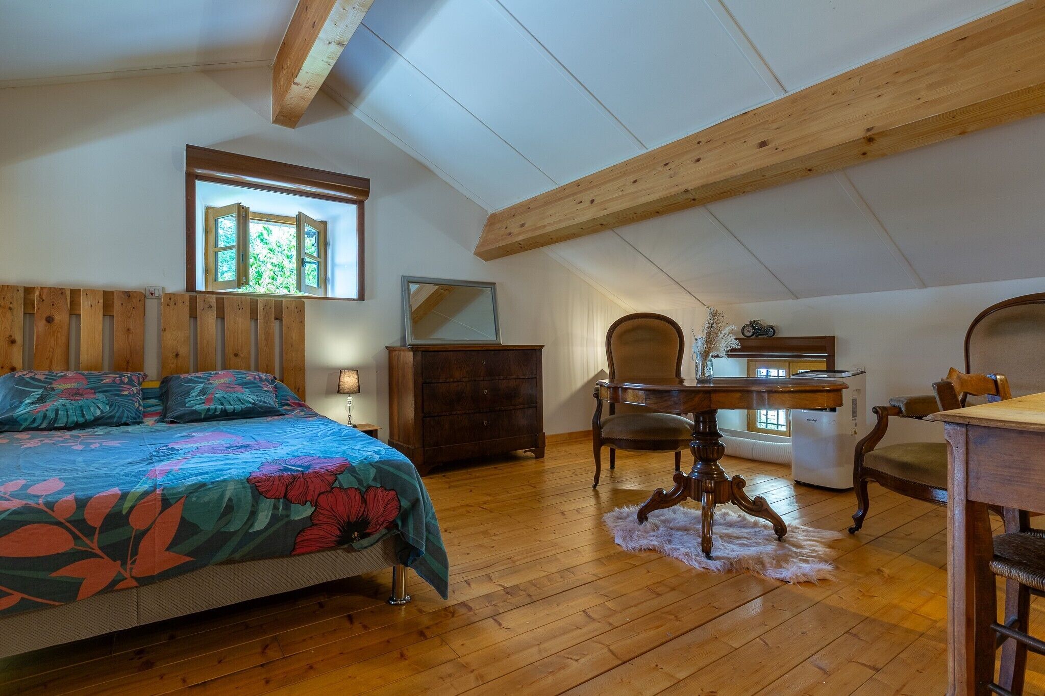 A bedroom with a bed, wooden ceiling beams, a dresser, a chair, a table, and a window.