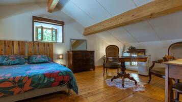 A bedroom with a bed, wooden ceiling beams, a dresser, a chair, a table, and a window.