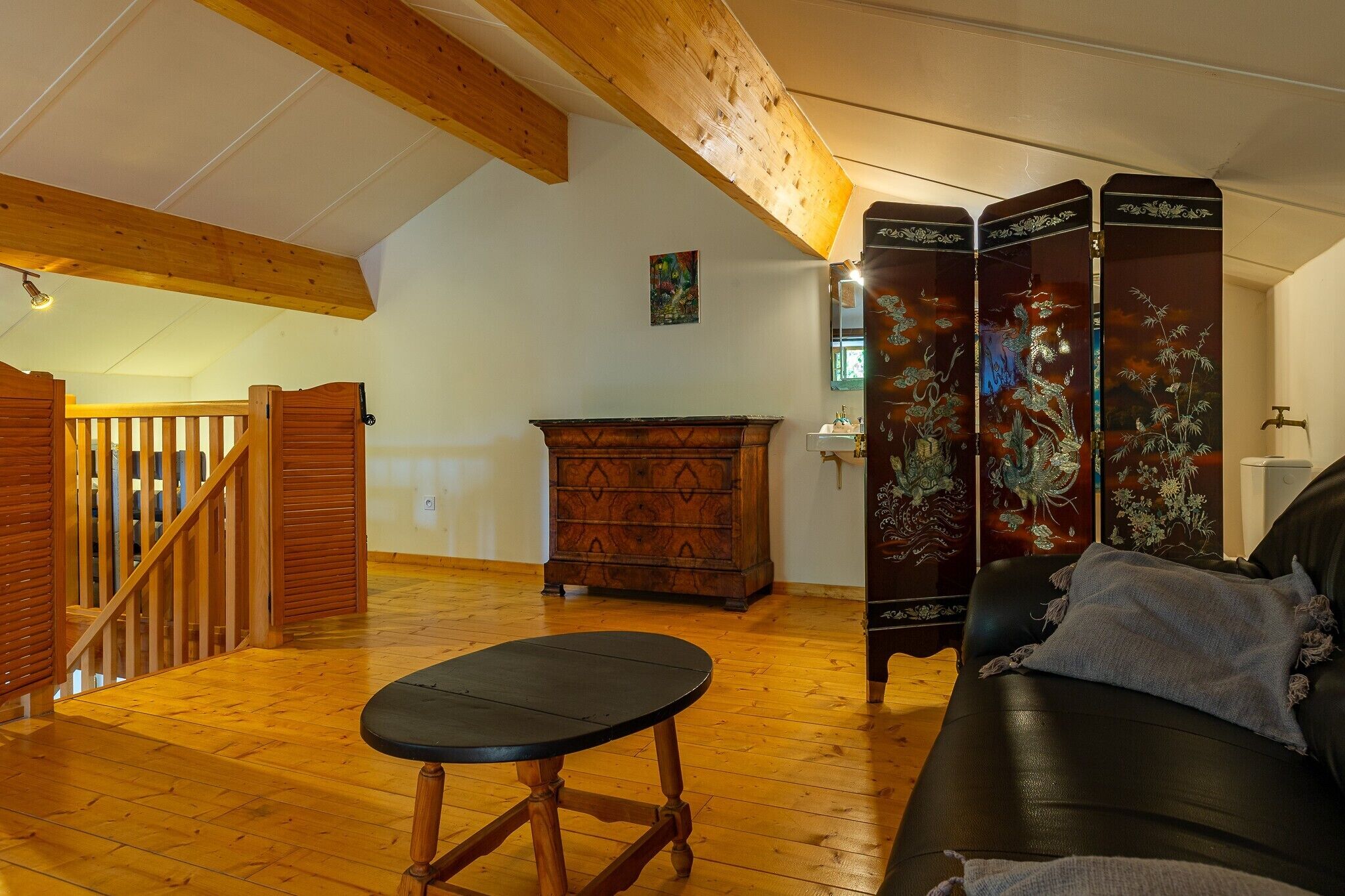 A room with a wooden staircase, a black leather sofa, a wooden cabinet, and a traditional decorative screen.