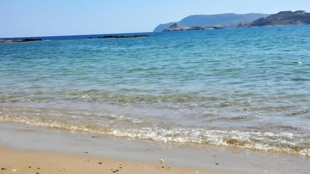 Ein Strand mit klarem Wasser, einem Sandufer und fernen Felsenformationen.