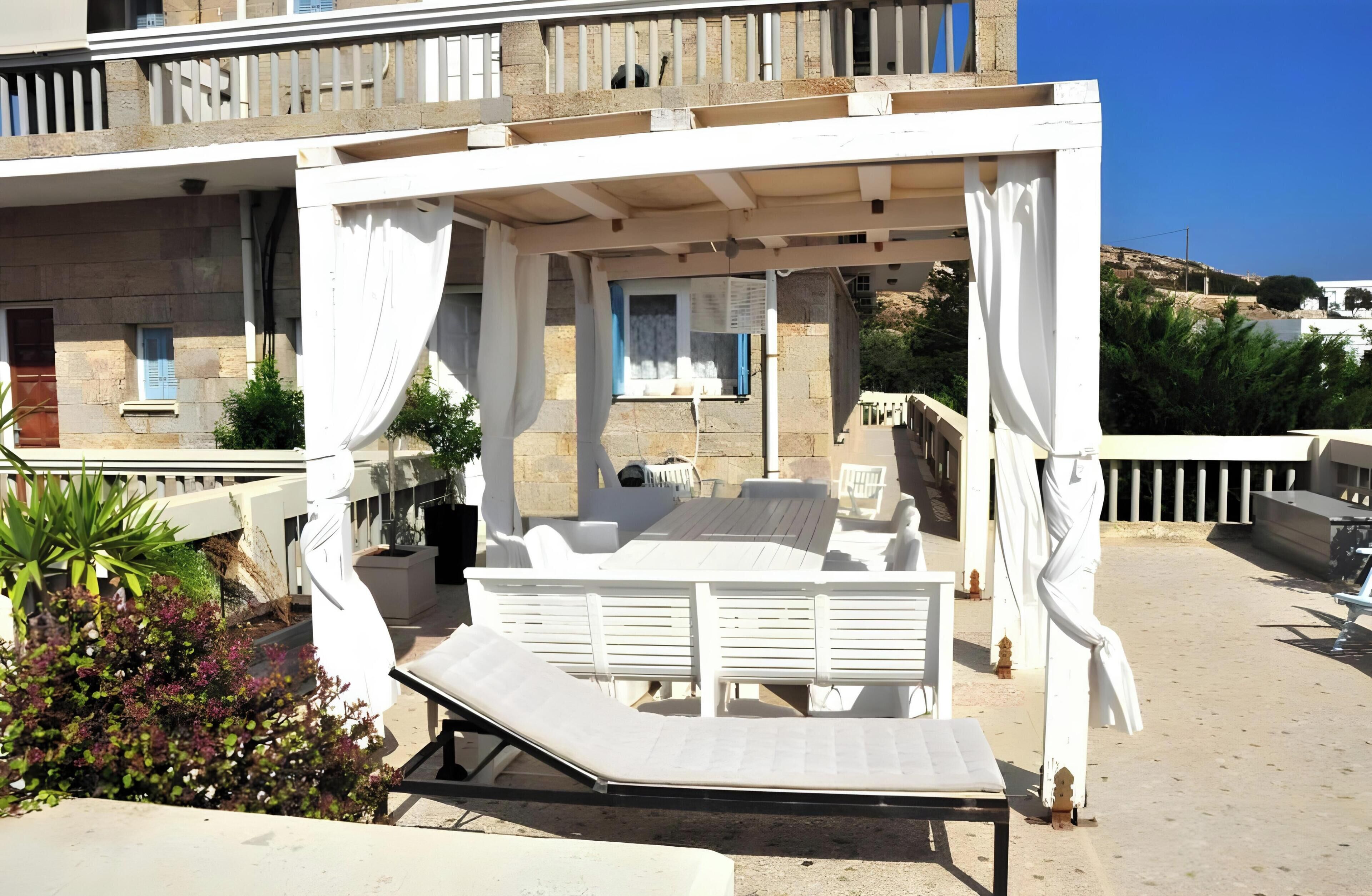 Ein überdachter Außenbereich mit Tisch und Stühlen, einem weißen Baldachin und Blick auf ein Gebäude mit Balkon.