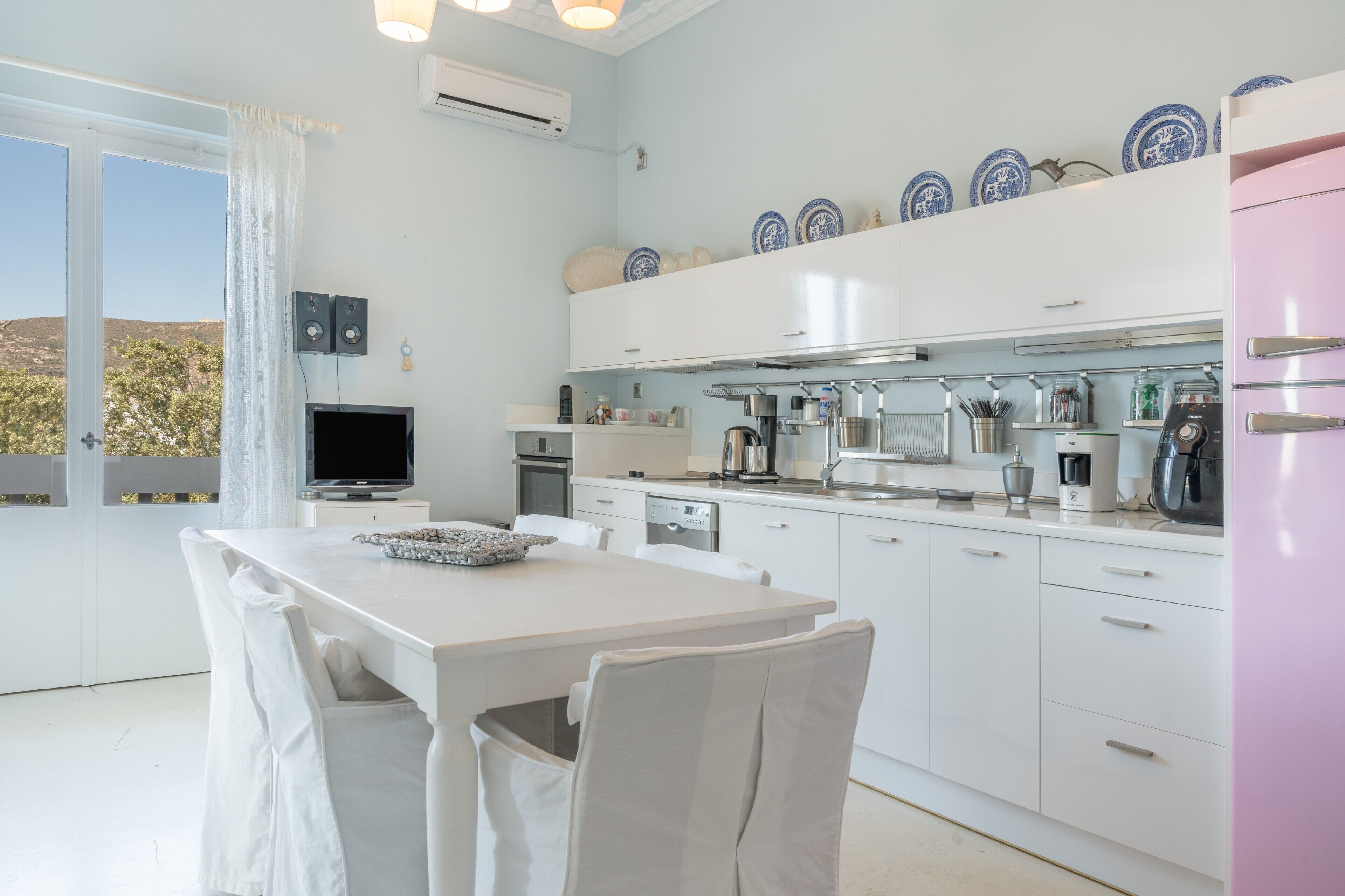 A modern kitchen with white cabinets, a dining table with chairs, a TV, and a view of the outdoors.
