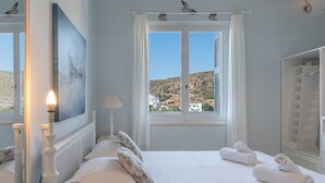 Ein Schlafzimmer mit Bett, Kissen, Nachttisch, Lampe, einem Fenster mit Vorhängen und Blick auf eine Hügelkuppe.