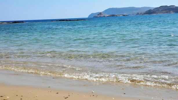 Ein Strand mit klarem Wasser, einem Sandufer und fernen Felsenformationen.