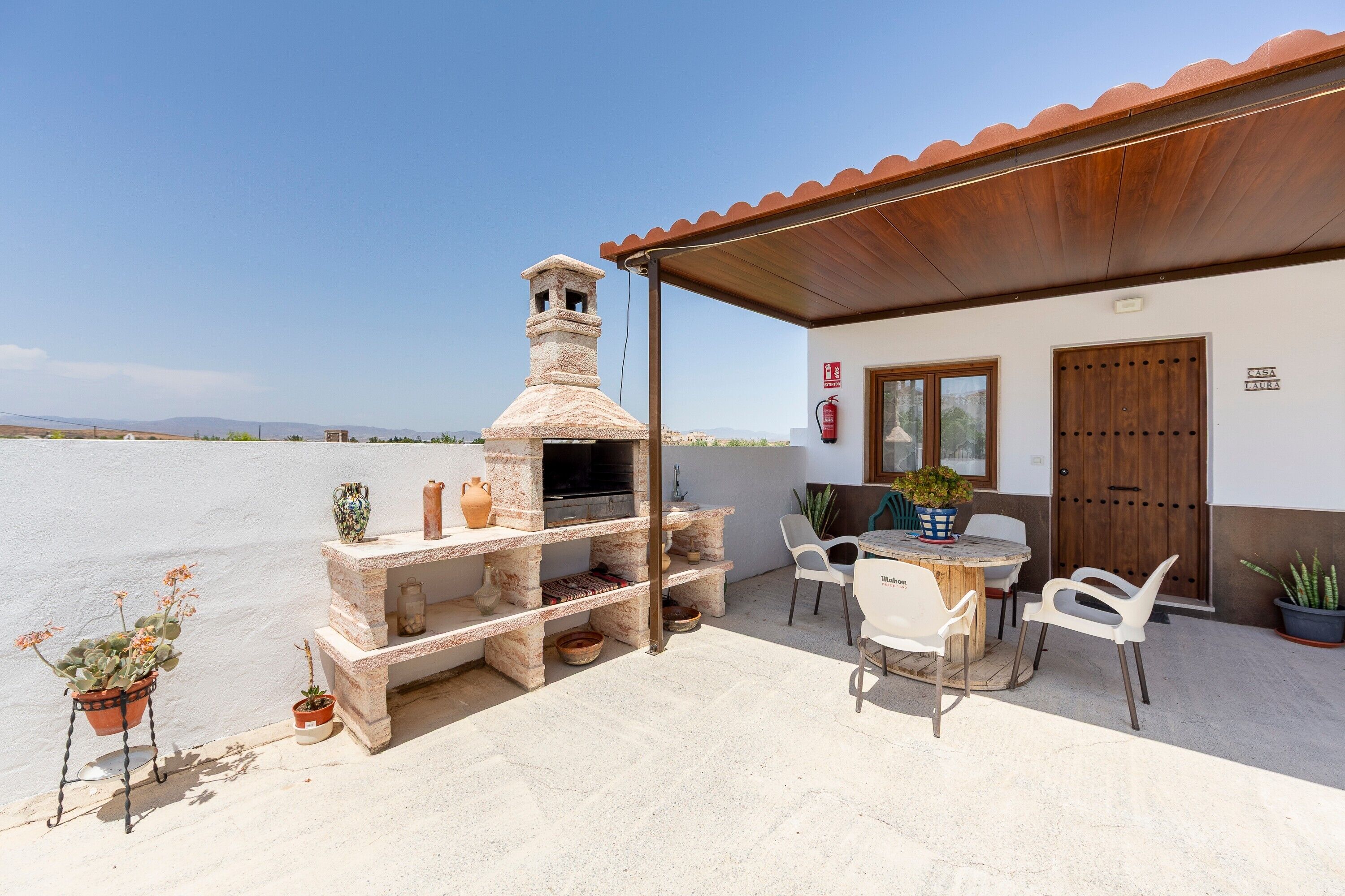 Un patio al aire libre con una barbacoa de piedra, sillas blancas, una mesa y plantas en macetas.