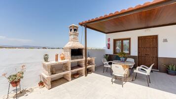 Un patio al aire libre con una barbacoa de piedra, sillas blancas, una mesa y plantas en macetas.