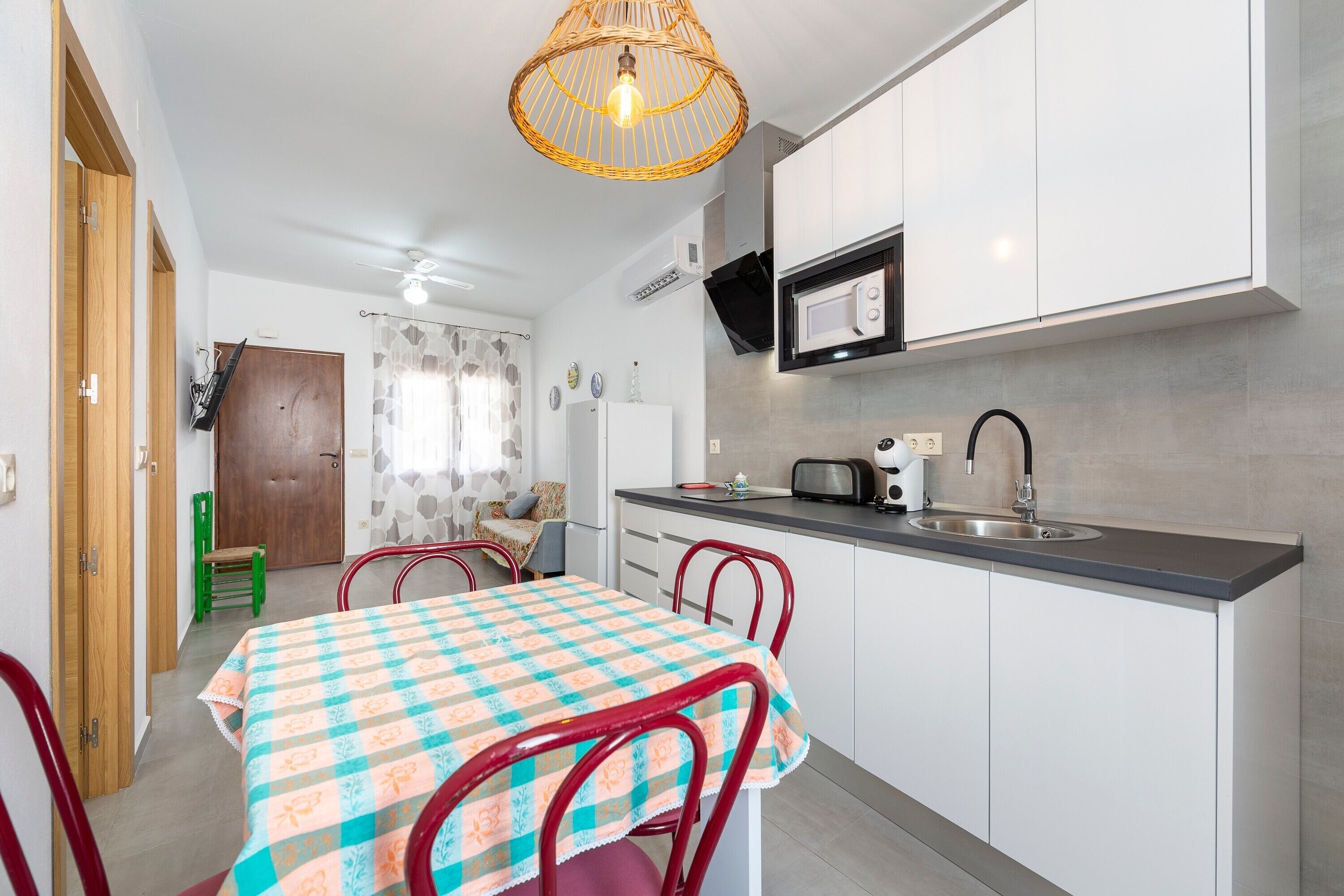 Cocina compacta con microondas, tostadora y lavamanos. Se aprecia una mesa de comedor con sillas y una lámpara de techo de mimbre.