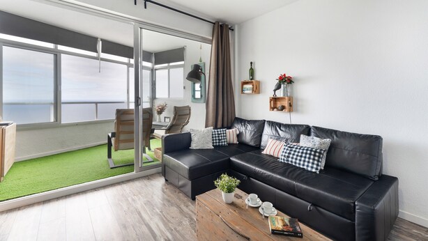 A modern living room with a black leather sofa, a wooden coffee table, and a view of a balcony with a green lawn. - 'The Blue Lighthouse' Studio with Sea View, Shared Pool, and Wi-Fi (La Matanza de Acentejo)