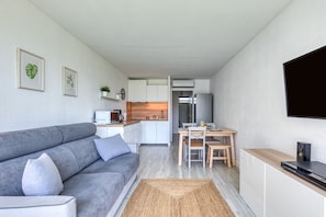 Una sala de estar moderna con un sofá gris, un televisor de pantalla plana montado en la pared y una zona de cocina con armarios blancos y encimera de madera.