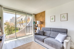 Una sala de estar moderna con un sofá gris, una estantería y un gran ventanal con vistas a edificios.