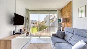Ein modernes Wohnzimmer mit Fernseher, grauem Sofa, Holzkonsole und Balkon mit Blick auf Gebäude und Grünflächen.