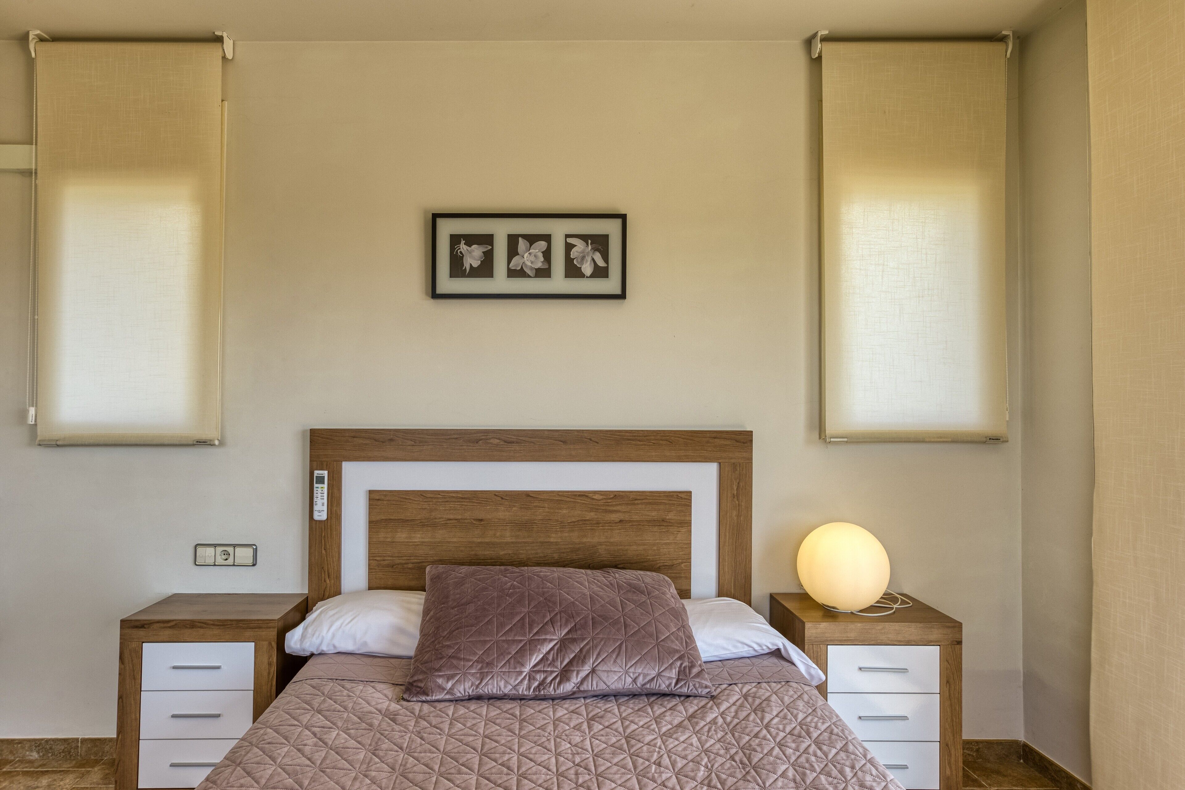 Een slaapkamer met een houten hoofdbord, een gewatteerde sprei, een bedlampje, een nachtkastje en een ingelijst kunstwerk aan de muur.