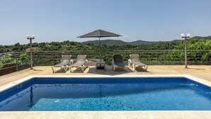 Une piscine entourée d’une clôture en métal et de verdure, avec des chaises longues et un parasol.