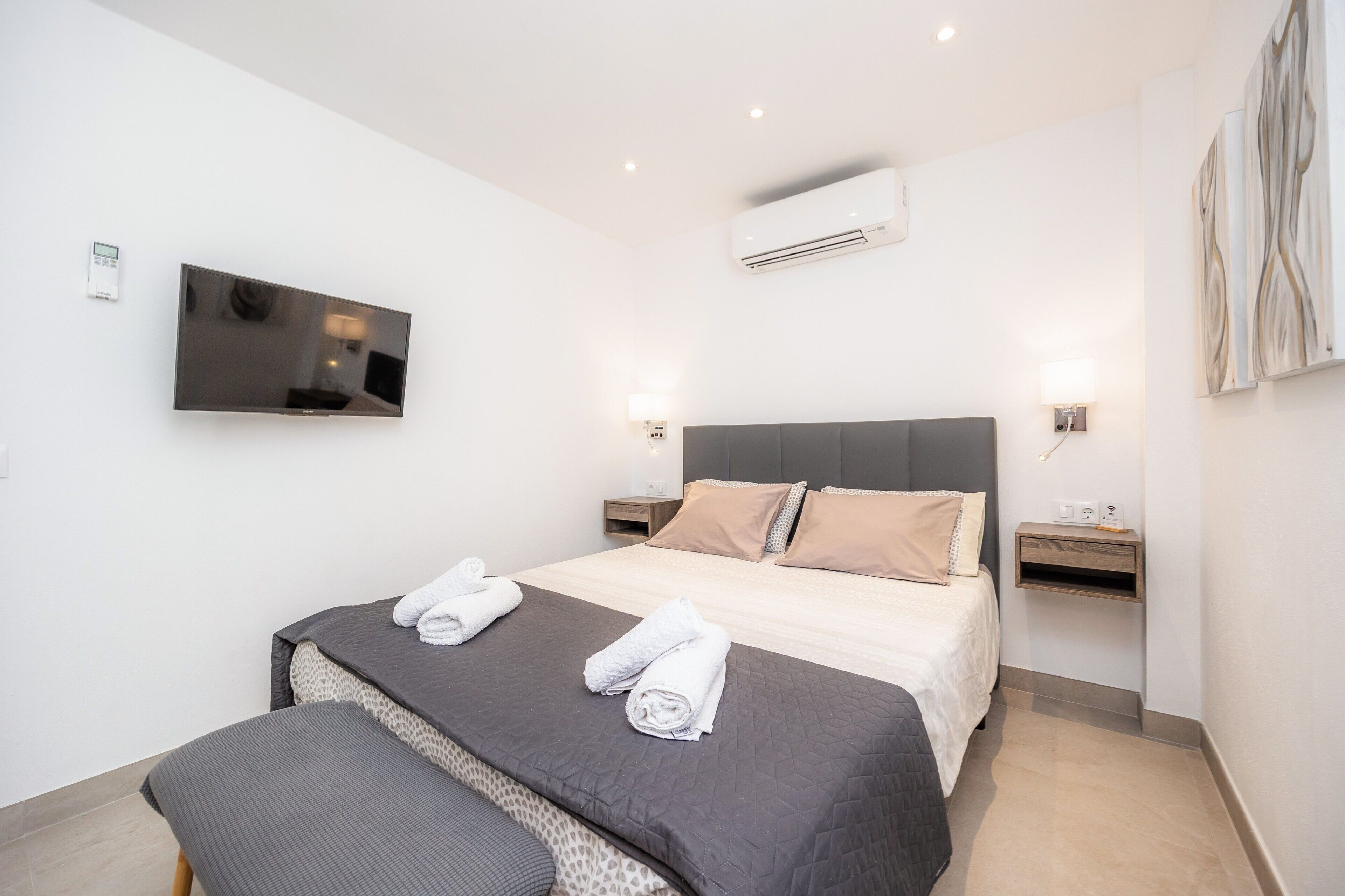 A modern hotel room with a large bed, bedside tables, a flat-screen TV, an air conditioning unit, and artwork on the wall.