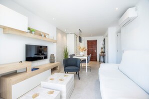 A modern living room with a flat-screen TV, a sofa, a dining area with chairs, and a wooden shelf.