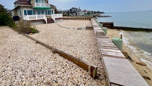 A beachfront property with a wooden walkway leading to a house with a balcony. - Relax and unwind at this 3-bedroom bayfront house in charming Mattituck. (Mattituck)