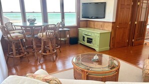 A living room with a view of the ocean, wicker furniture, a green cabinet, and a television on the wall. - Relax and unwind at this 3-bedroom bayfront house in charming Mattituck. (Mattituck)
