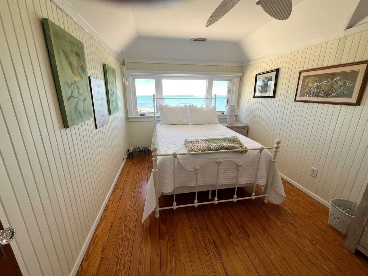 A bedroom with a white bed, wooden flooring, and a view of the sea through a window.
