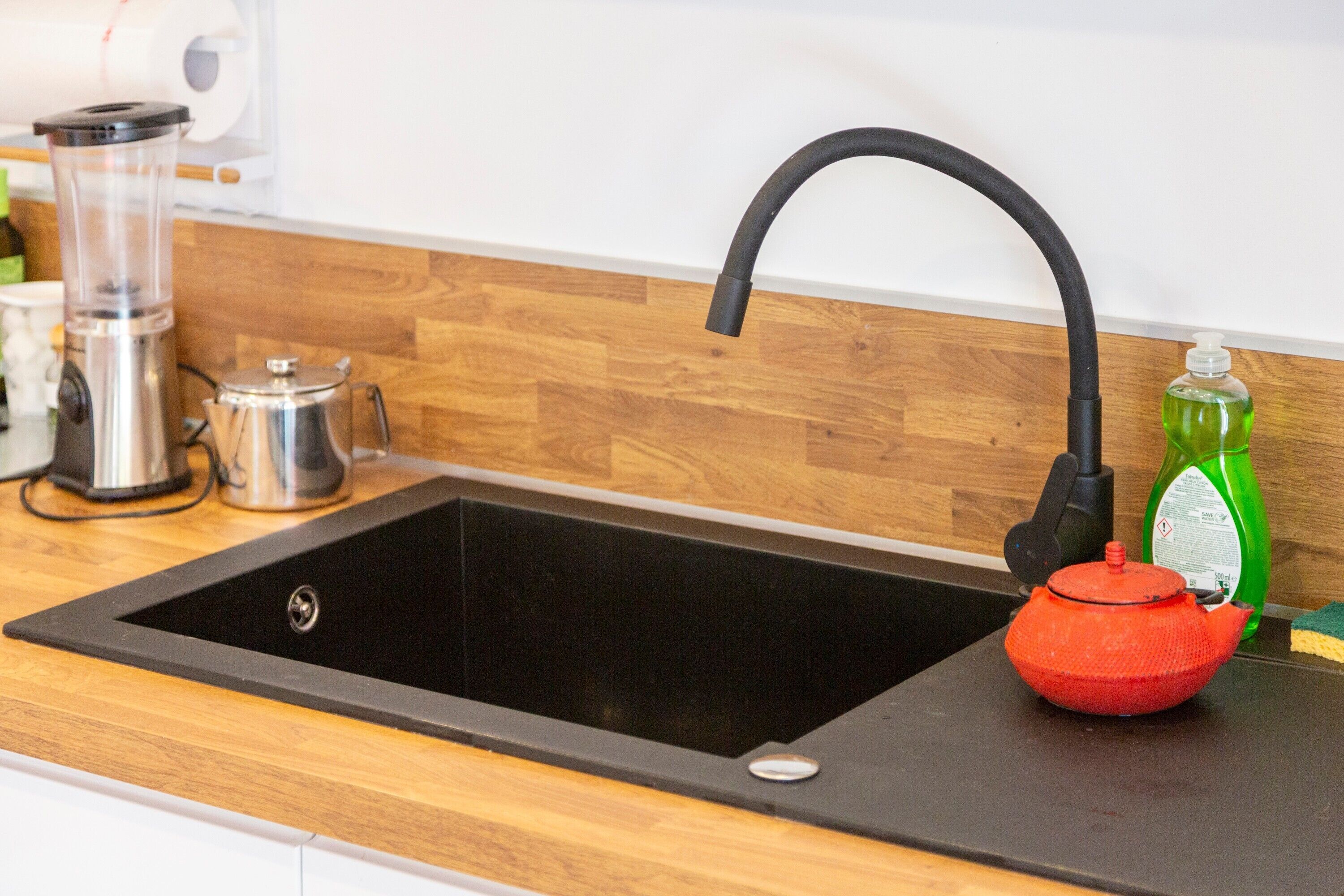 A kitchen sink with a black faucet, a red teapot, and a green bottle of dish soap.