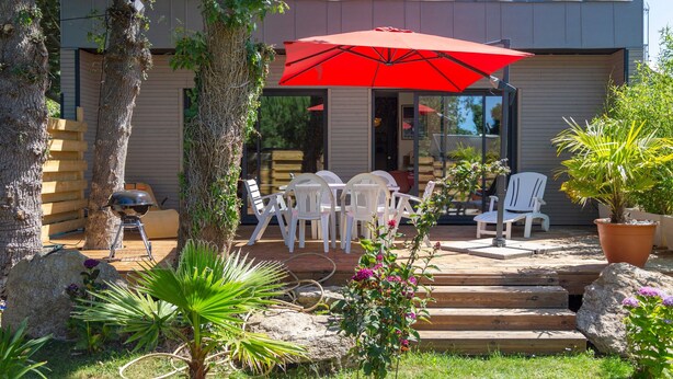 Un patio exterior moderno con una sombrilla roja, sillas blancas y una parrilla para barbacoa.