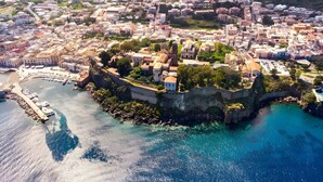 Vista aérea de un pueblo costero con una imponente estructura fortificada, un puerto deportivo y una zona urbana densa con edificios.