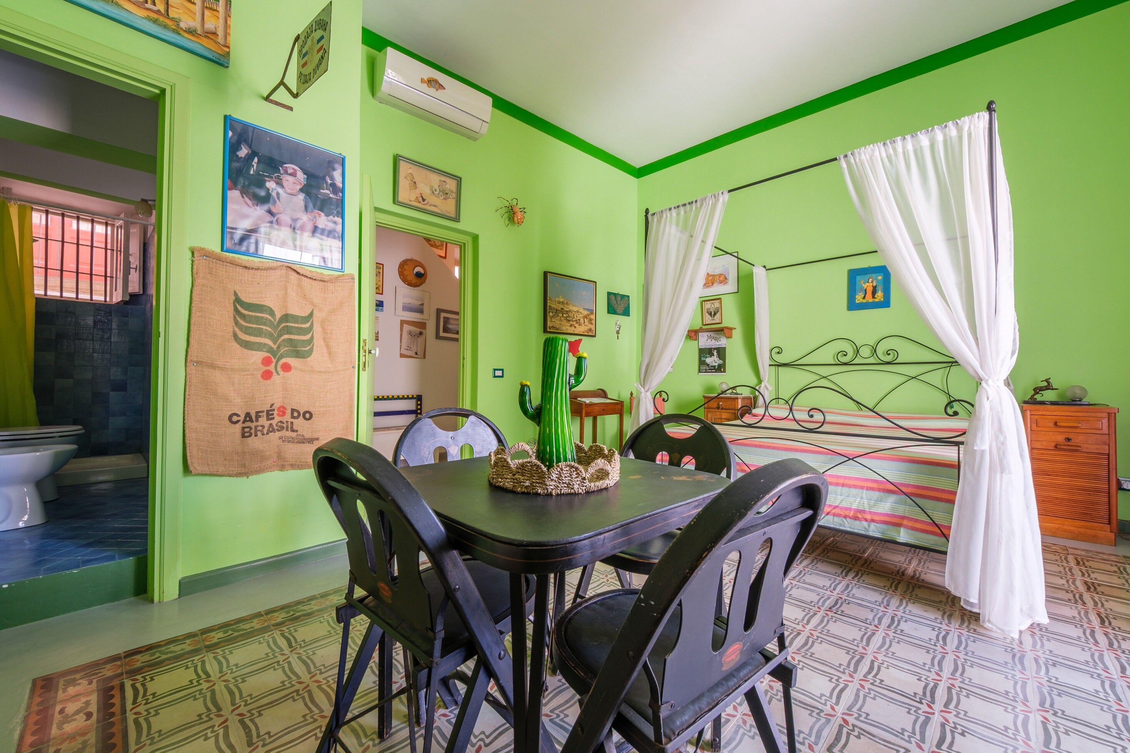 Una habitación con cama, una mesa con sillas, un ventanal con cortinas, y una pared con cuadros y una toalla colgada.