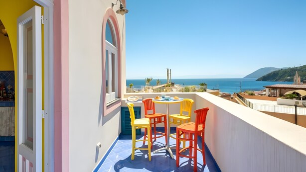 Un balcón con mesa y sillas, vista al mar y un edificio con fachada rosa y amarilla.