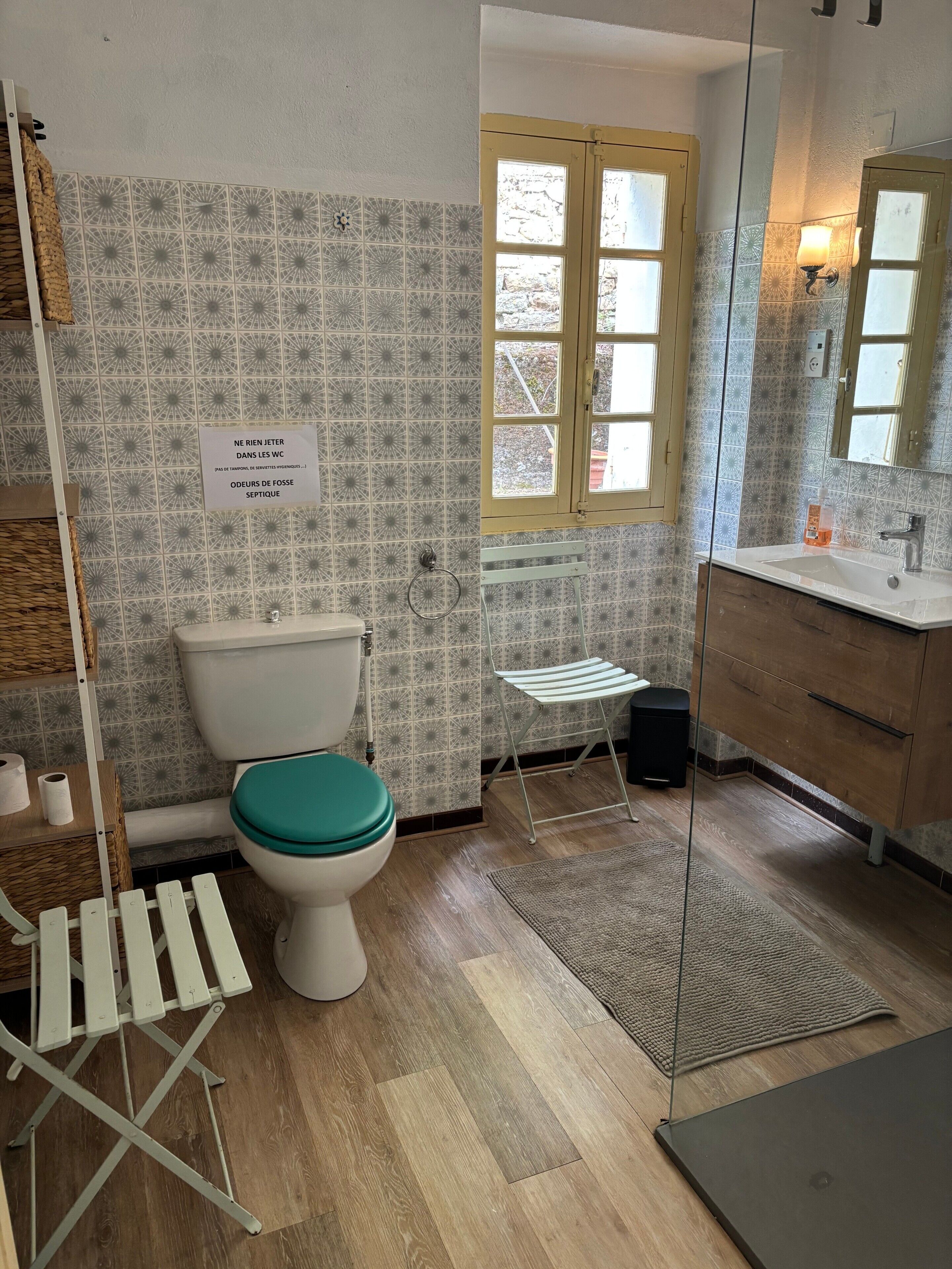 Un bagno con piastrelle decorate, un water con lo sgabello verde acqua, un lavandino con specchio e una finestra con vista.