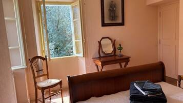 A bedroom with a bed, a chair, a desk with a mirror, and a window with a view of trees.
