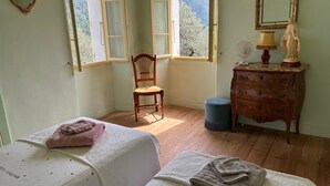 Une chambre avec deux lits, une chaise en bois, une commode, un miroir et une fenêtre donnant sur des arbres.
