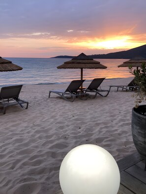 Une plage avec des chaises longues et des parasols, un lampadaire sphérique blanc et une vue sur la mer au coucher du soleil.