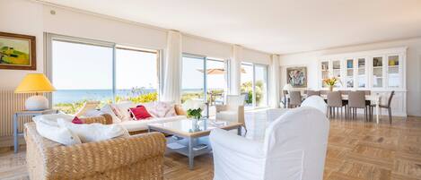 Amplia sala de estar con un ventanal grande con vistas al mar, zona de comedor con un aparador blanco y una acogedora disposición de asientos.