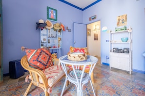 Una stanza con una sedia in vimini, un tavolino rotondo, una porta blu e uno scaffale bianco con diversi oggetti.