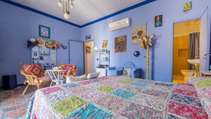 A room with a blue wall, a bed with a colorful quilt, a chair with a cushion, a lamp, and a door leading to a bathroom. - Room 'Blu' with Shared Terrace, Wi-Fi and Air Conditioning (Lipari)