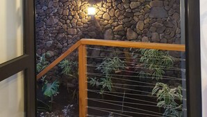 Une vue à travers une porte vitrée donnant sur une pièce avec un mur en pierre, des poutres en bois au plafond et une rambarde en métal ornée de plantes.