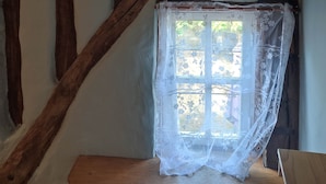 A room with exposed wooden beams, a small window with a lace curtain, and a wooden bench.