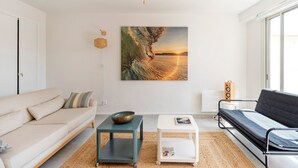 A modern living room with a sofa, a small table, a bean bag, a wall-mounted speaker, and a large painting of a sunset over water. - Apartment 'Notre Dame' with Sea View, Private Terrace and Wi-Fi (Capbreton)