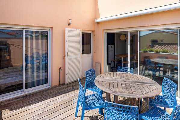 A wooden deck with a round table and blue chairs, a sliding glass door, and a view of the interior. - Apartment 'Notre Dame' with Sea View, Private Terrace and Wi-Fi (Capbreton)