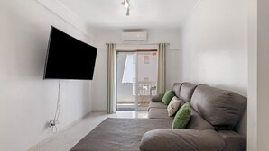 A living room with a flat-screen TV mounted on the wall, a grey sofa with green cushions, and a sliding glass door leading to an outdoor area. - Apartment 'Excellent  In Olhos D´Água' with Balcony, Wi-Fi and Air Conditioning (Olhos de Água)