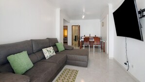 A living room with a sectional sofa, a flat-screen TV mounted on the wall, and a dining area with a table and chairs. - Apartment 'Excellent  In Olhos D´Água' with Balcony, Wi-Fi and Air Conditioning (Olhos de Água)
