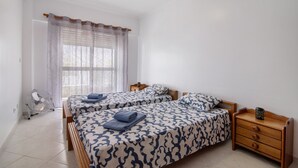 A bedroom with two beds, a bedside table, a nightstand, a fan, and a window with curtains. - Apartment 'Excellent  In Olhos D´Água' with Balcony, Wi-Fi and Air Conditioning (Olhos de Água)
