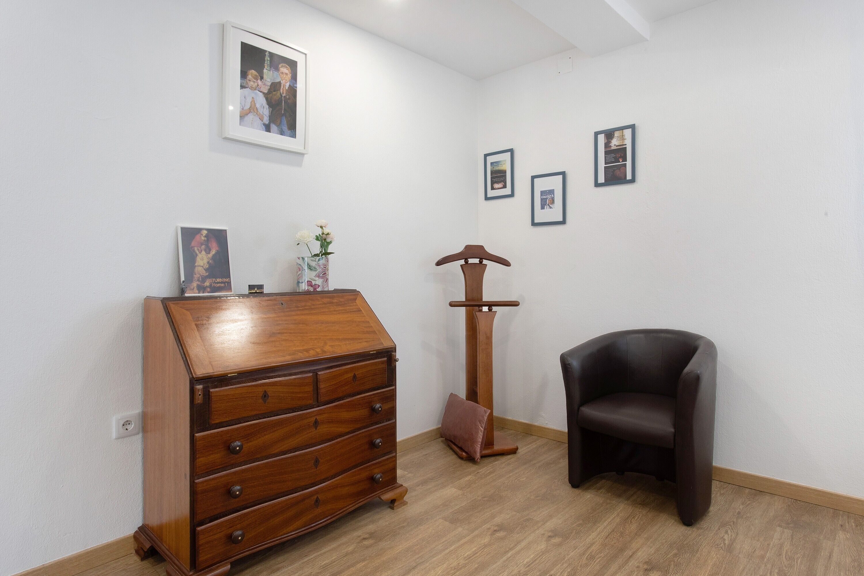 A room with a wooden dresser, a black leather chair, a tall floor lamp, and framed pictures on the wall.