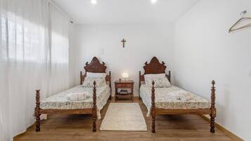 Un dormitorio con dos camas de madera, una mesita de noche, una cruz en la pared y una ventana con cortinas.