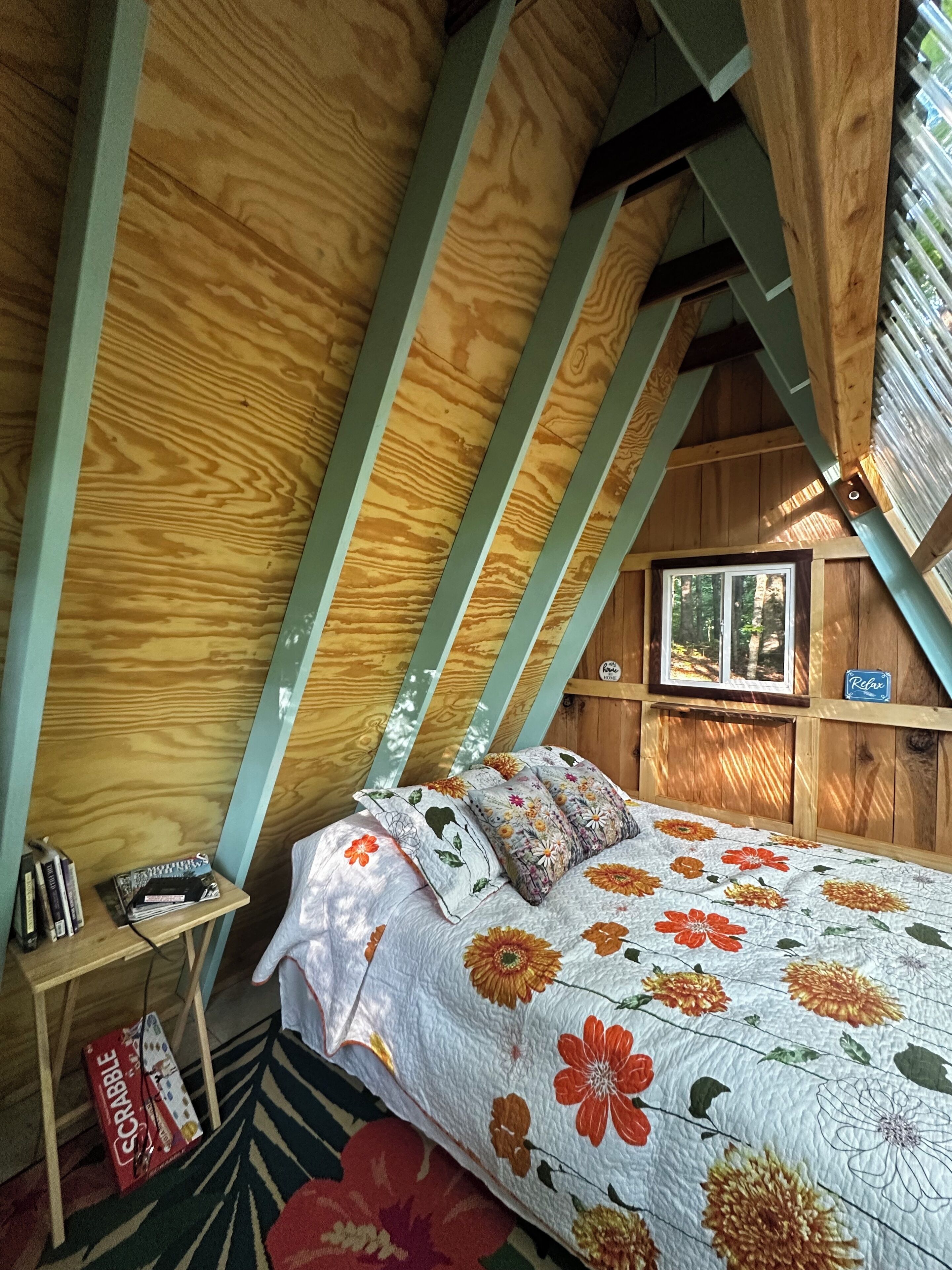 Een gezellig slaapkamer onder de dakpannen met een houten schuine hemel, een bed met bloemenbedekking, een kleine tafel met boeken en een gezelschapsspel, en een raam met uitzicht op bomen.