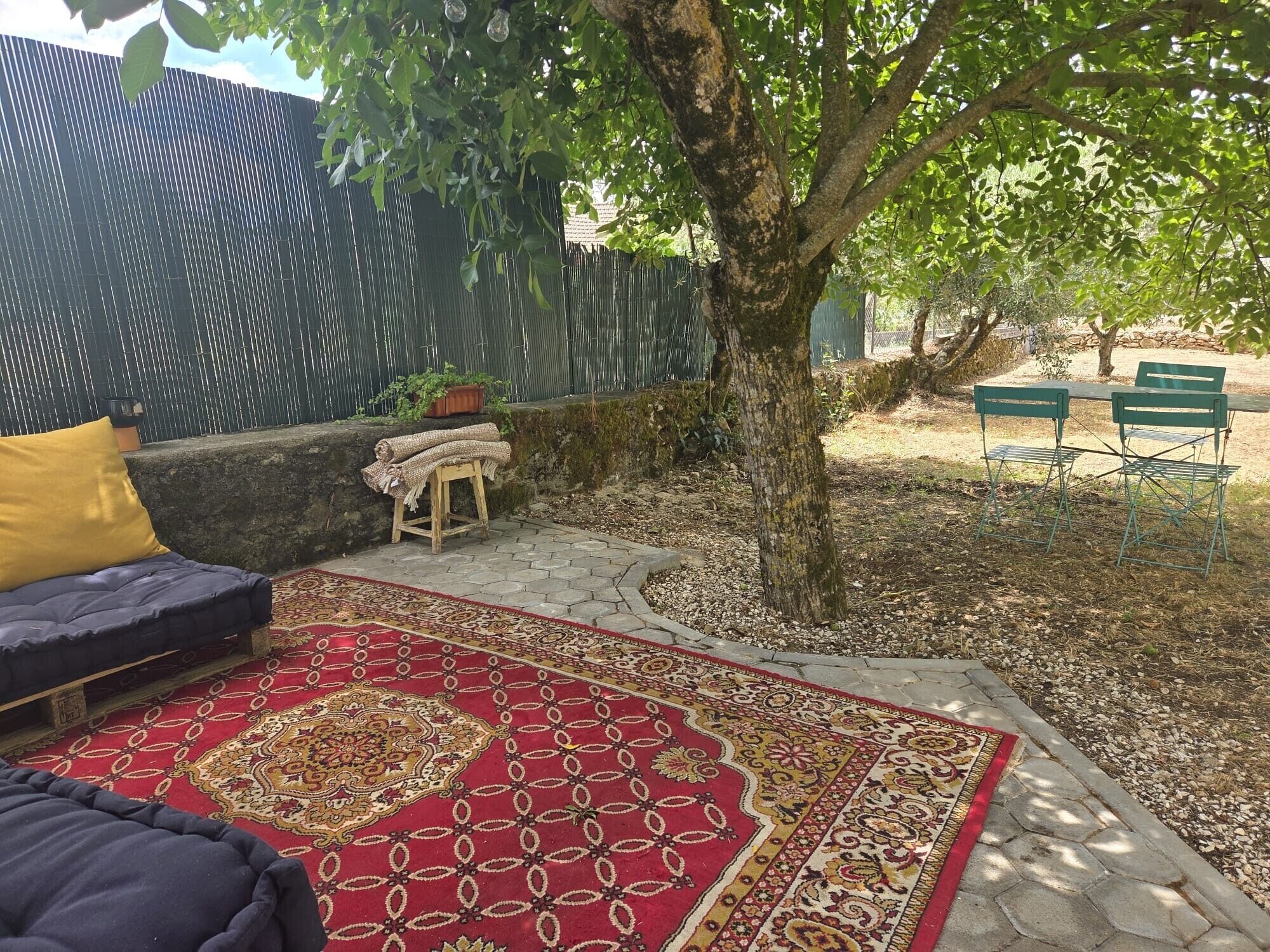 Un patio con alfombra estampada, un sofá con cojín amarillo, un taburete pequeño con una manta y un árbol.